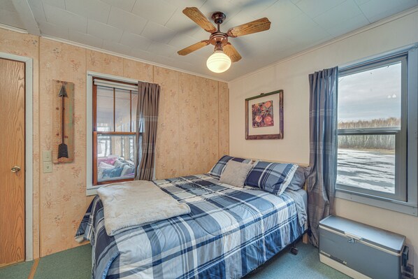 Un dormitorio con cama, ventilador de techo, ventana con cortinas, un cuadro en la pared y un mueble pequeño.