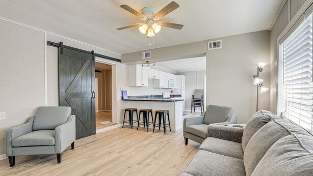 A modern living room with a large gray sofa, two armchairs, and a wooden floor. A kitchen with white cabinets and a sliding barn door is visible in the background. - Sun-soaked Okeechobee Retreat w/ Canal Views! (Okeechobee)