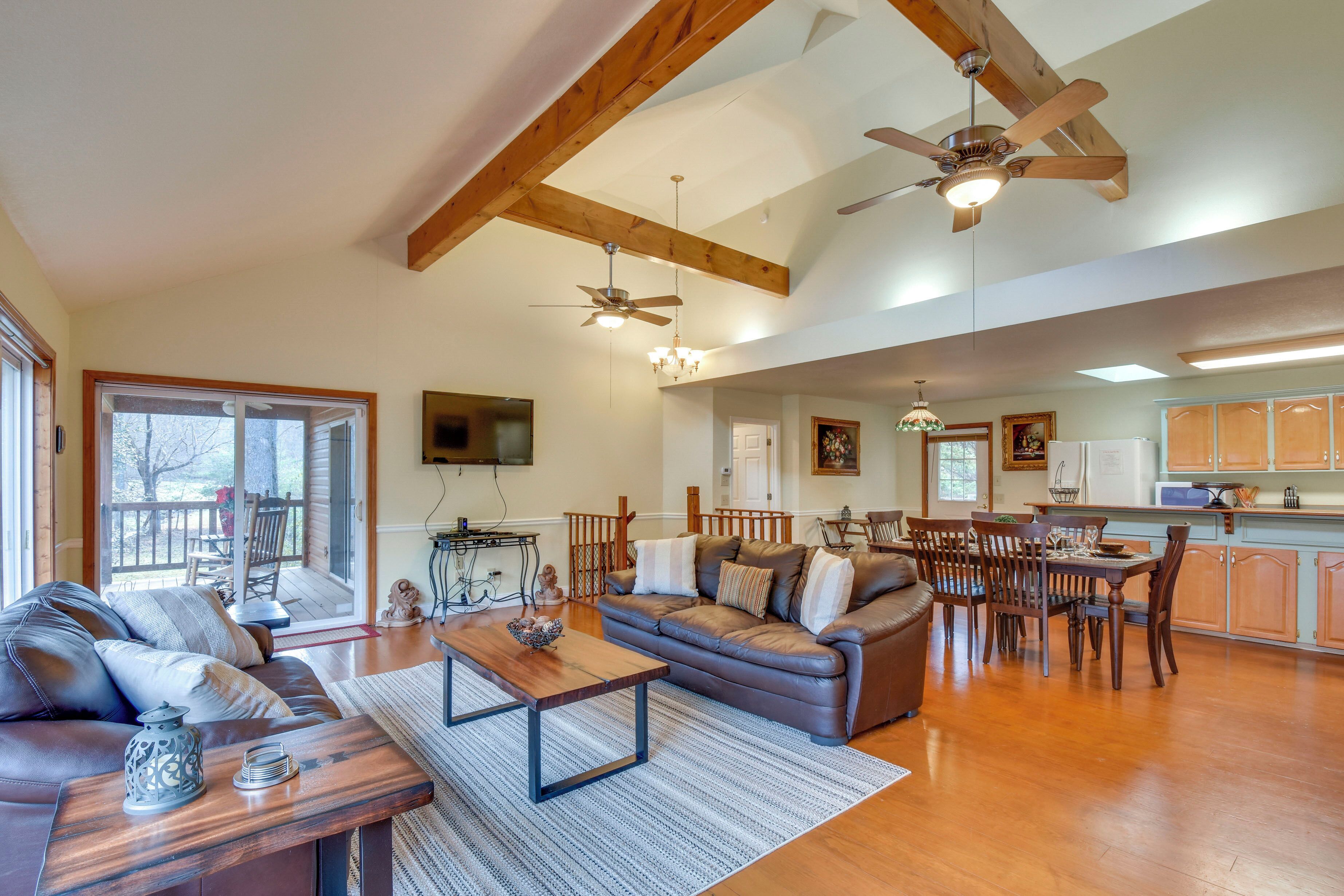 A spacious living room with a large sofa, a wooden coffee table, and a dining area with a table and chairs. There is a ceiling fan, a chandelier, and a kitchen area visible in the background.