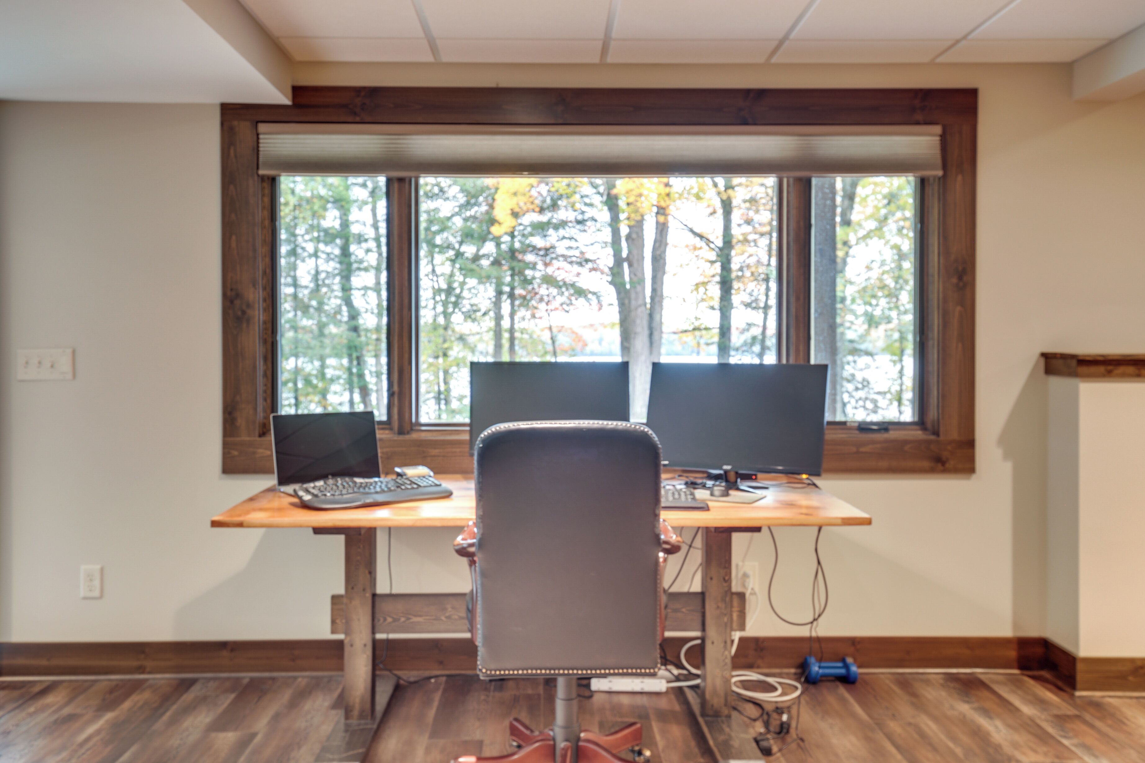 Una habitación con un escritorio de madera, dos computadoras portátiles, una silla y una ventana que da a un paisaje de árboles.