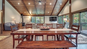 Un comedor amplio con una mesa y sillas de madera, un ventilador de techo y ventanas grandes que ofrecen vistas al exterior.
