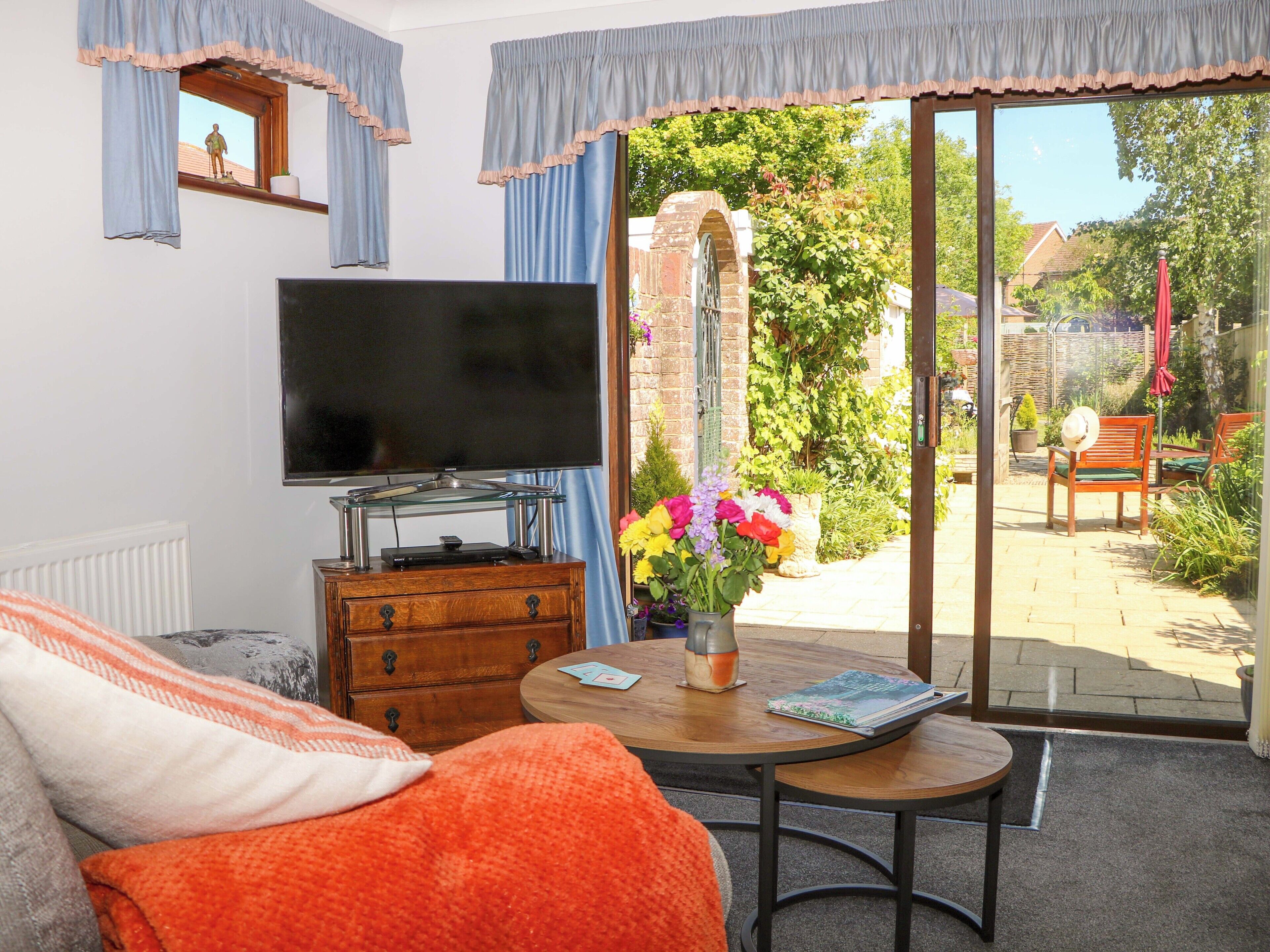 A living room with a television, a wooden dresser, a coffee table with a vase of flowers, and a view of the garden through a glass door.