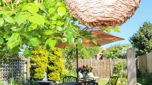 Een terras buiten met een geweven parasol, stenen zitplaatsen, en een tuin met planten en een houten omheining.