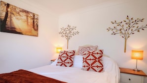 A neatly made bed with a brown blanket, two patterned pillows, a wall-mounted painting of a forest, and two wall-mounted lamps. - Lavender Cottage (Hailsham)