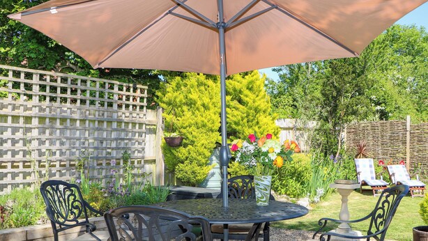 Een terras buiten met een ronde tafel en vier stoelen, een grote parasol en een decoratieve vaas met bloemen.