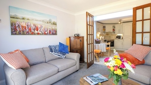 A living room with a grey sofa, a coffee table with a book and a vase of flowers, and a painting of a field on the wall. - Lavender Cottage (Hailsham)