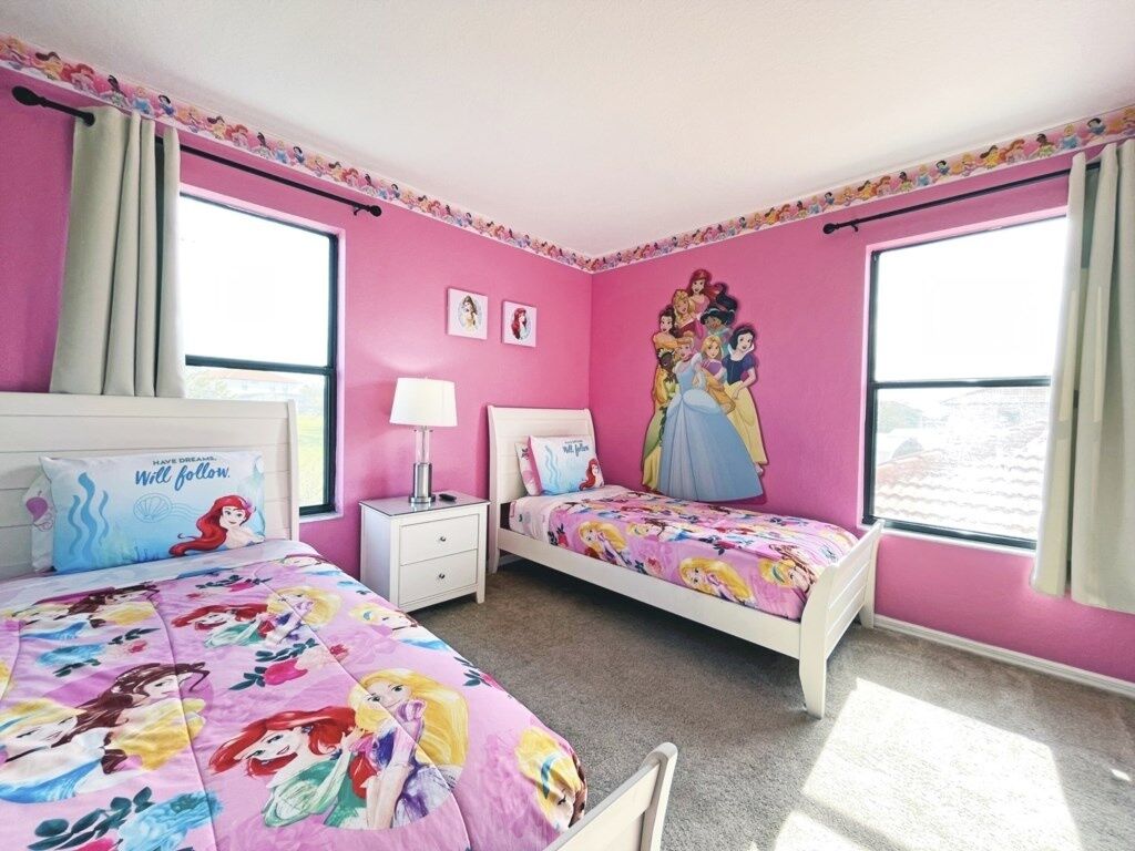 A bedroom with pink walls, two beds with Disney princess bedding, a nightstand, a lamp, and a window with curtains.
