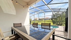Un espace extérieur abrité, aménagé pour les repas, avec une table et des chaises en verre, une terrasse en briques et une vue sur une piscine et des maisons en arrière-plan.