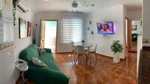 Ein Wohnzimmer mit einem grünen Sofa, einem Esstisch mit Stühlen, einem Fernseher an der Wand und einer Zimmerpflanze.
