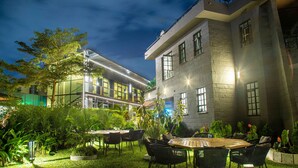 Un espace extérieur aménagé en terrasse, avec une table et des chaises, entouré de verdure et bordé d’un bâtiment doté de grandes fenêtres.