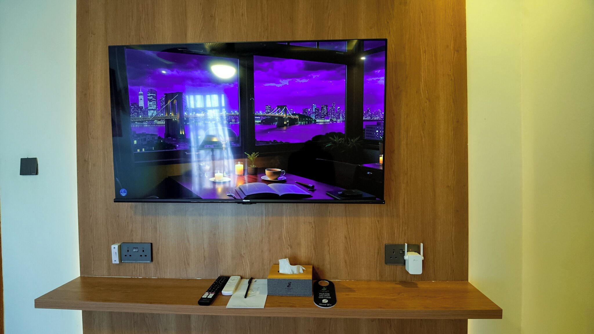 Uma TV de tela plana fixada numa parede de madeira, um console de madeira abaixo com um controle remoto, um livro, uma caixa de lenços e um carregador de celular.