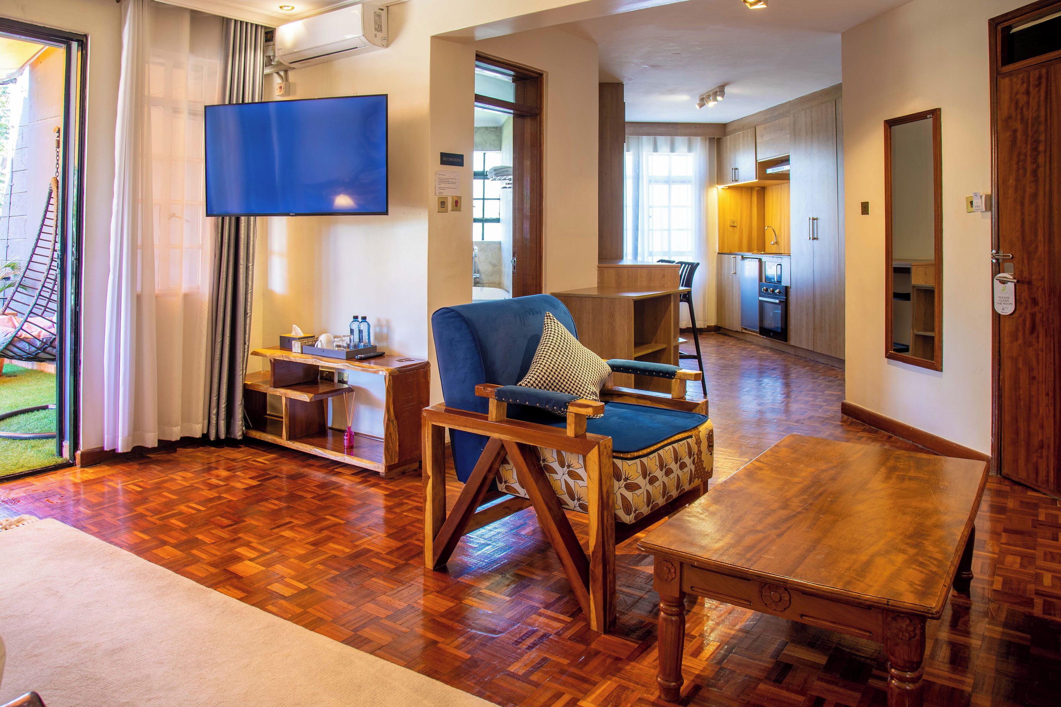 A hotel room with a wooden floor, a blue armchair, a wooden table, a flat-screen TV, and a kitchen area.
