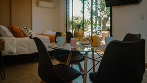 Moderni hotellihuone, jossa on sänky, ruokapöytä tuoleineen, seinälle asennettu televisio ja suuri ikkuna, josta avautuu näkymä puita kohti.