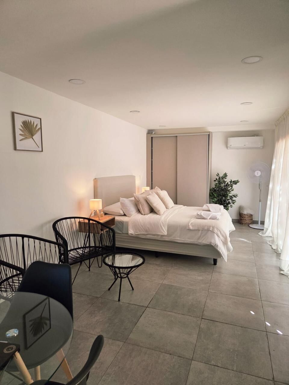 A modern hotel room with a bed, bedside table, lamp, chairs, and a potted plant.