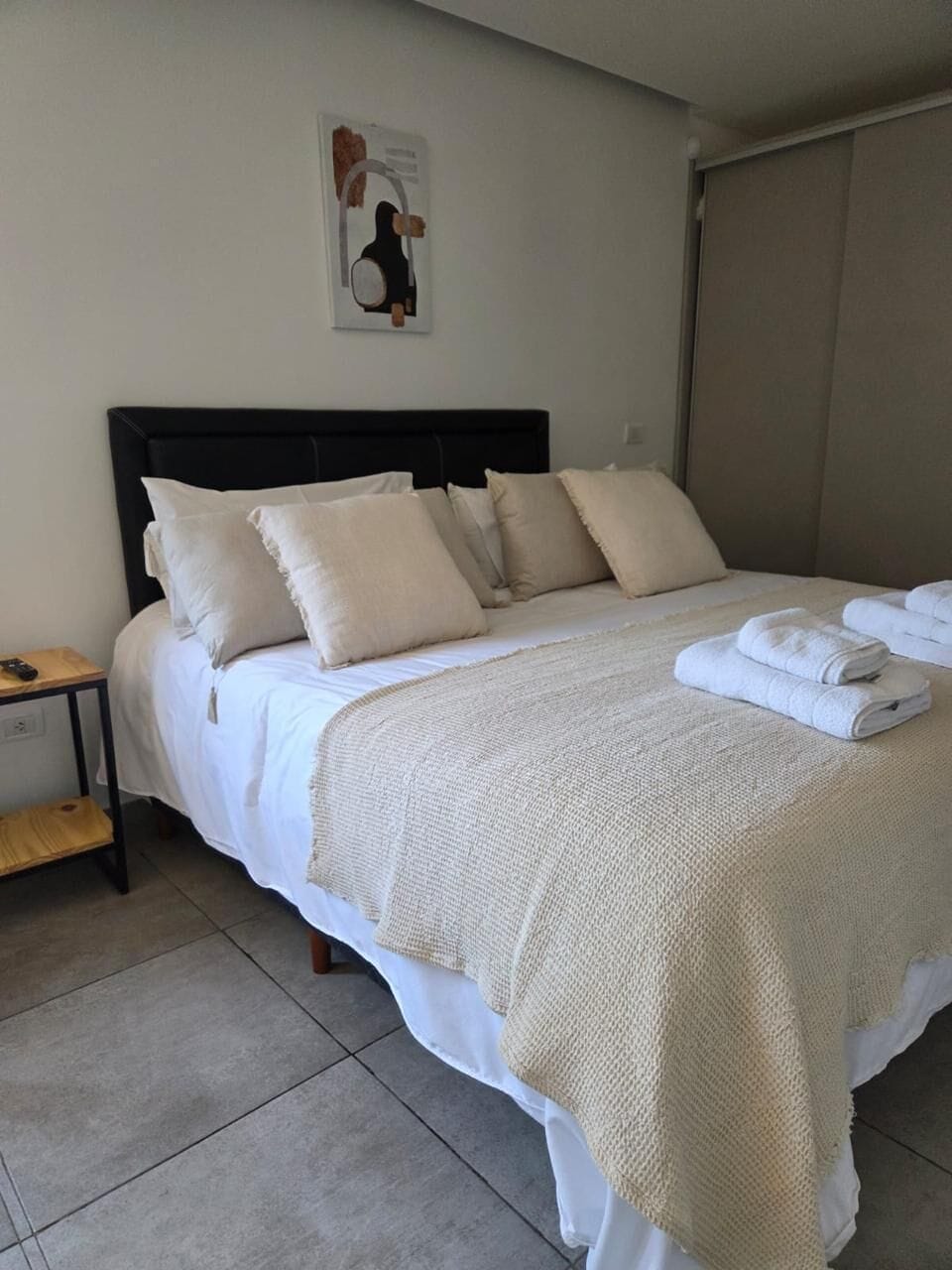 A neatly made bed with a white comforter and pillows, a folded towel on the bed, and a small bedside table with a lamp.