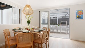 A dining area with a round table set for six, surrounded by wicker chairs, a hanging light fixture, and a view of the outdoors through a glass door. - Coastal Escape – waterfront reserve on Jervis Bay (Vincentia)