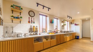 A modern kitchen with wooden cabinets, white countertops, and various kitchen appliances. There are shelves with plants and decorative items, and a clock on the wall. - Bloom Hotel - Ranchi (Ranchi)