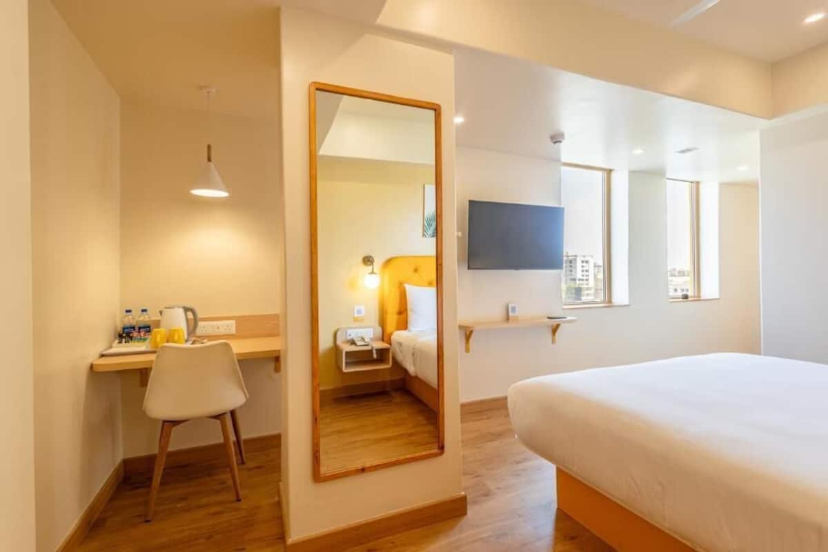 A hotel room with a bed, a desk with a chair, a mirror, a TV mounted on the wall, and a window.