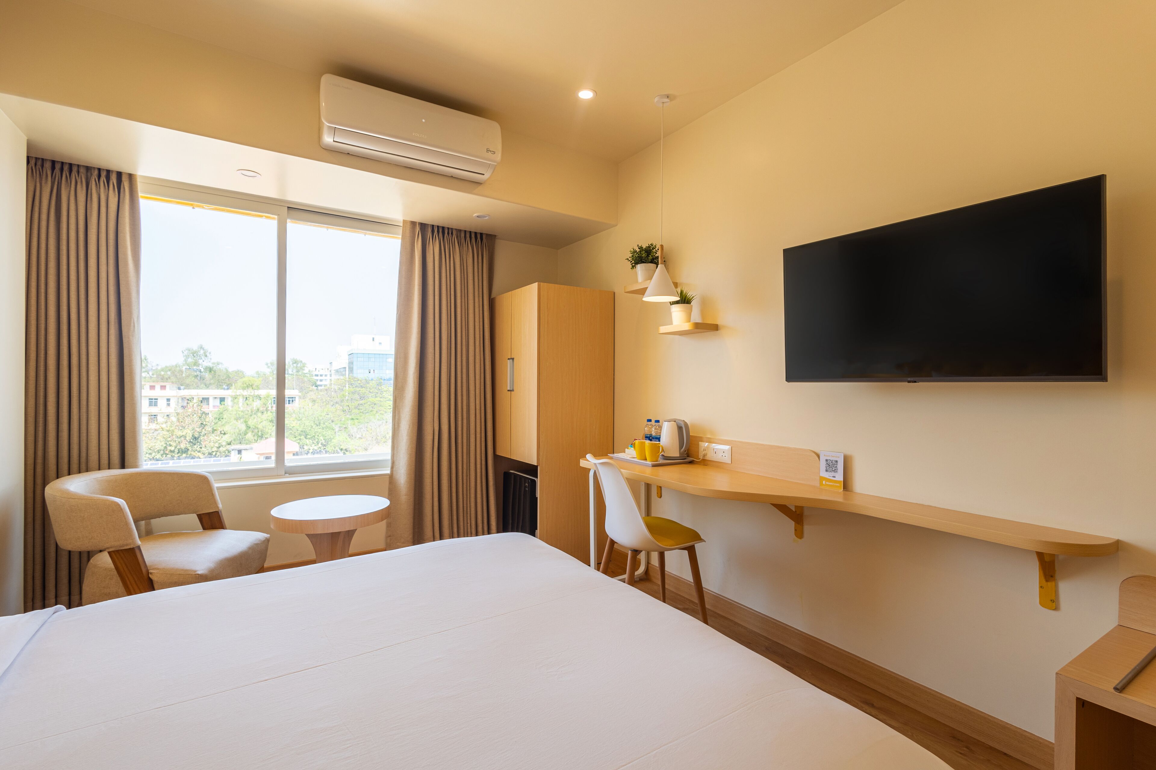 A hotel room with a large bed, a desk with a chair, a TV mounted on the wall, and a window with curtains.