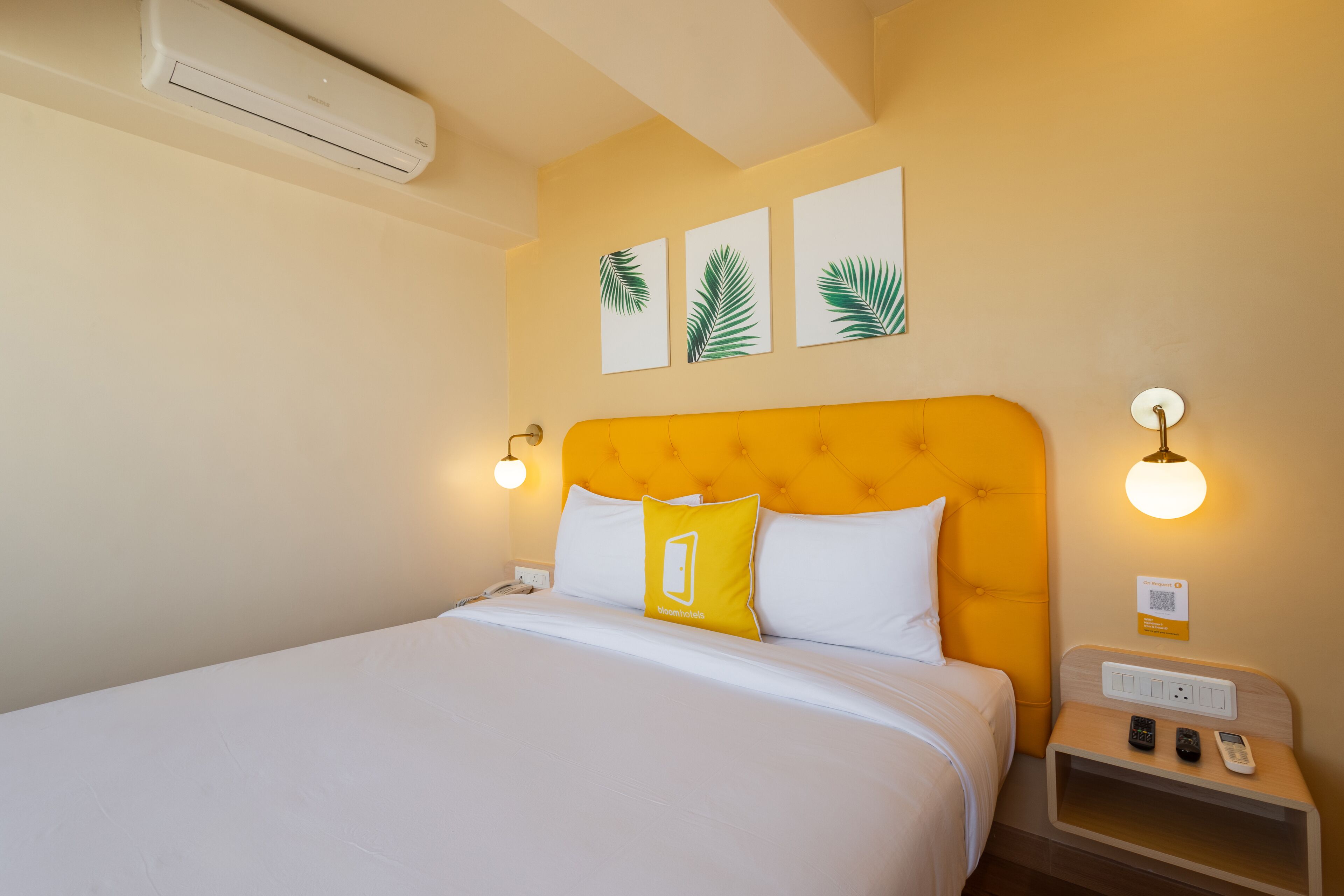 Une chambre d’hôtel comprenant un lit, une table de chevet, un climatiseur fixé au mur et trois tableaux représentant des motifs de feuilles.
