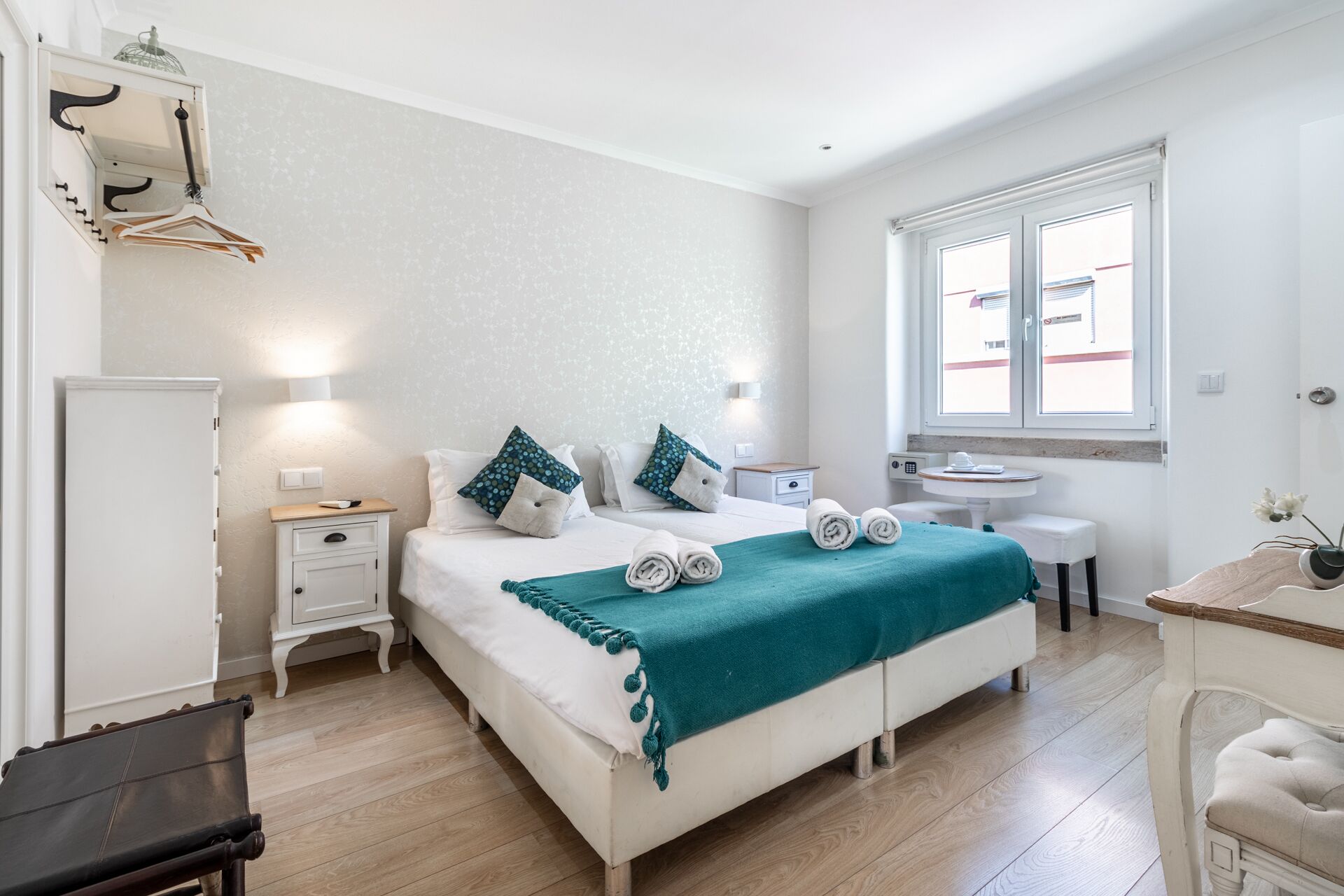 Un dormitorio ordenado con cama, mesitas de noche, una pequeña mesa y una ventana.