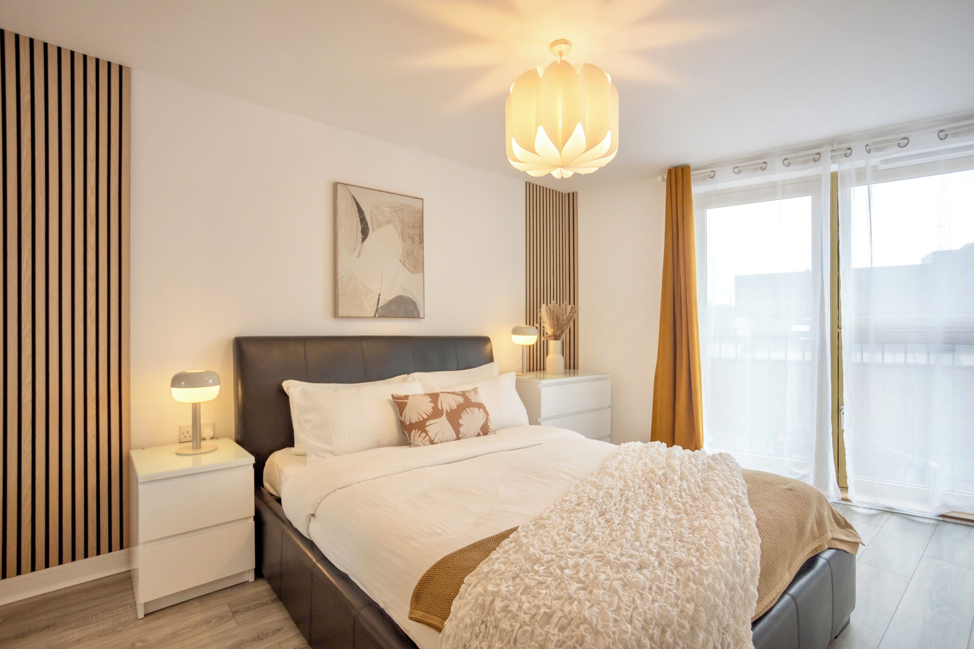 A modern bedroom with a large bed, bedside tables with lamps, a ceiling light, a painting on the wall, and a view through the window.
