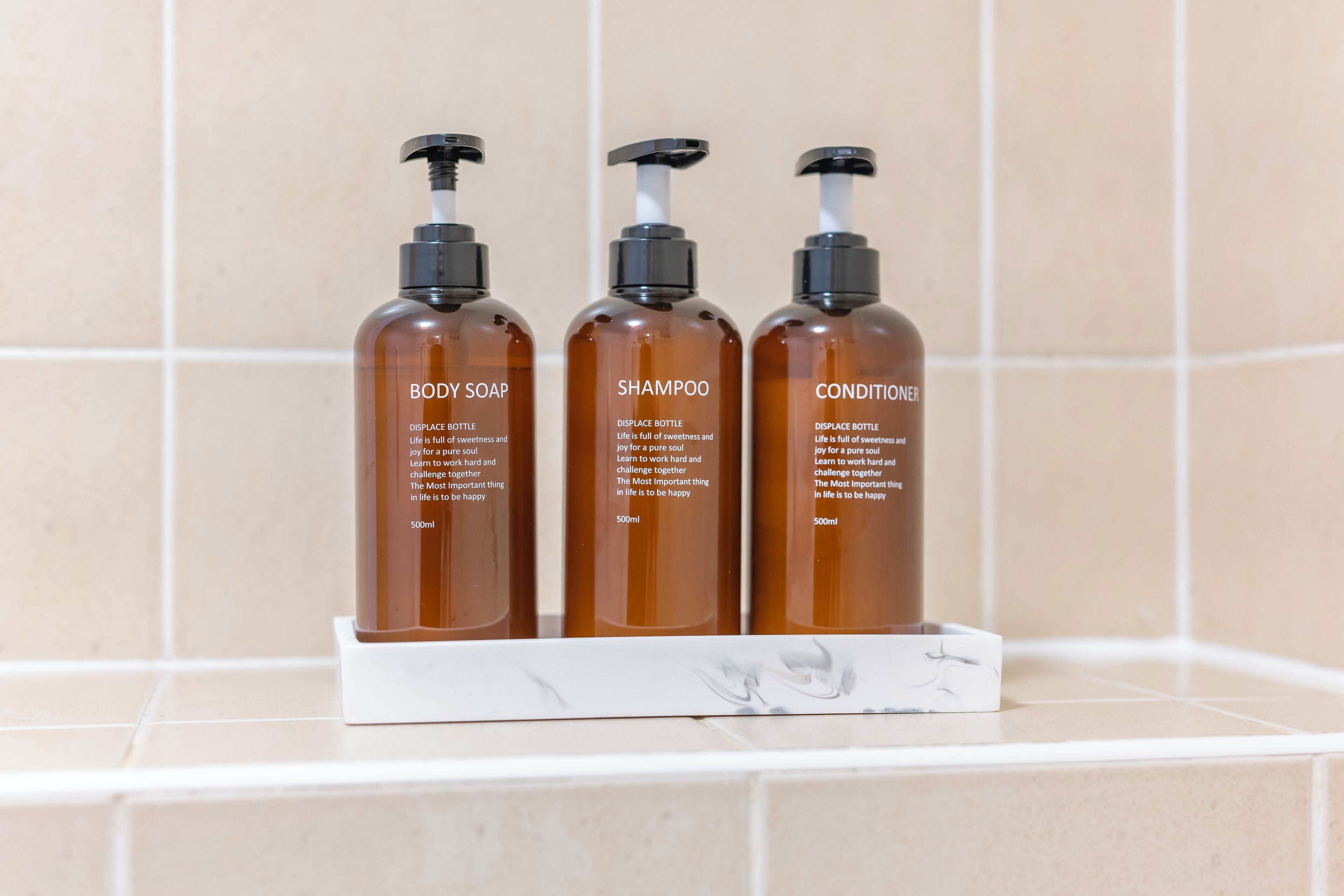 Three bottles of body soap, shampoo, and conditioner on a white marble shelf against a tiled wall.