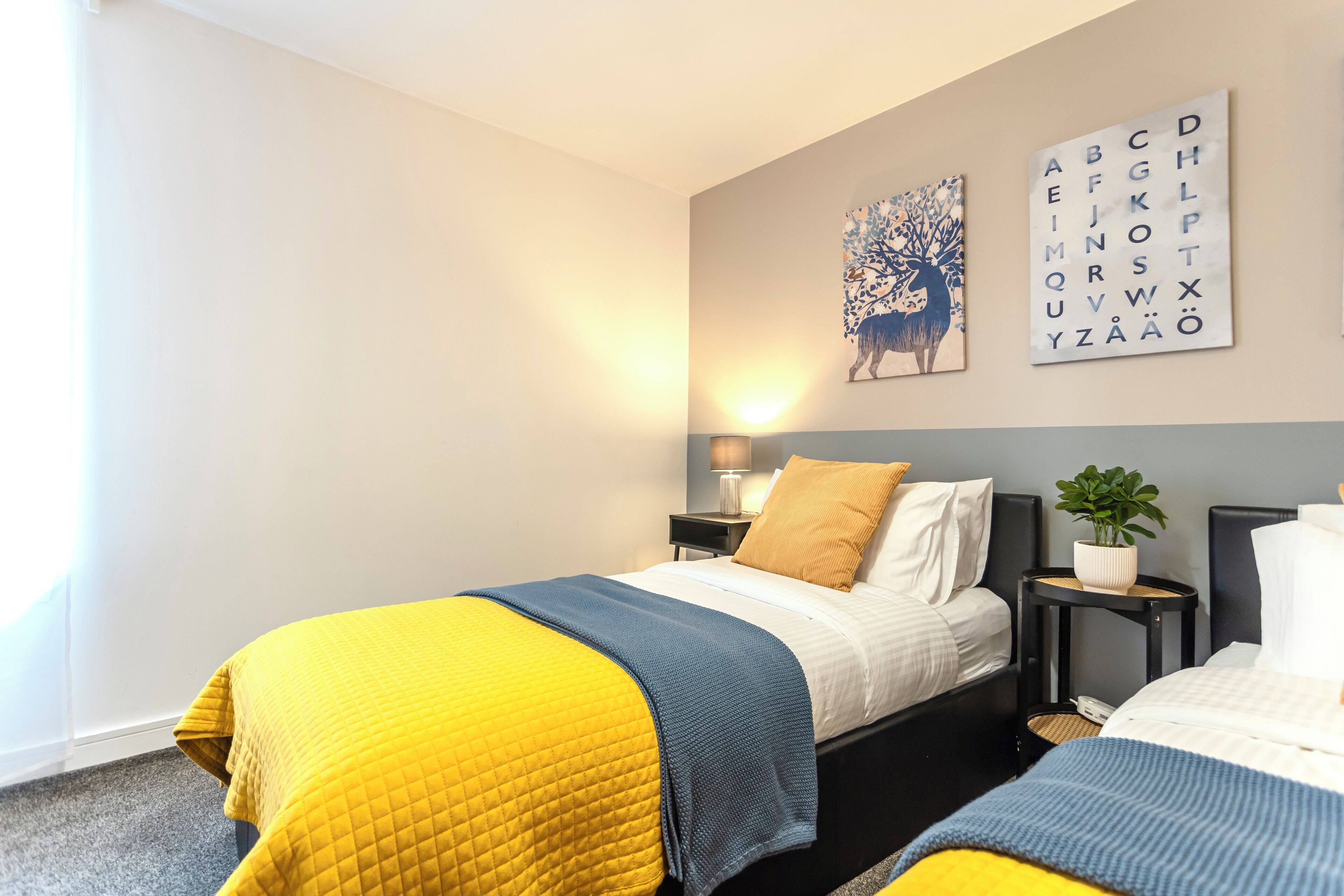 Une chambre d’hôtel moderne avec deux lits, une table de chevet avec une lampe et une œuvre d’art représentant l’alphabet, accrochée au mur.