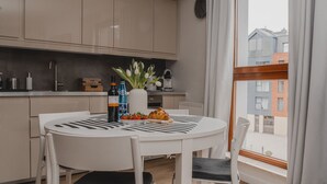 A modern kitchen with white cabinets, a round dining table with chairs, a window with curtains, and a view of buildings outside. - Waterlane Island Gdańsk Apartment 15 (Sopot)
