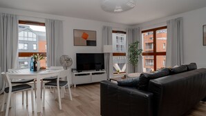 A modern living room with a black leather sofa, a dining table with chairs, a flat-screen TV, and large windows with curtains. - Waterlane Island Gdańsk Apartment 15 (Sopot)