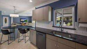 Cocina moderna con encimera negra, lavamanos de acero inoxidable y armarios de madera clara. Hay una zona de comedor con dos sillas negras y una mesita. Un ventanal ofrece vistas al exterior.