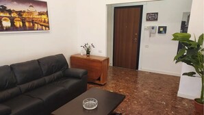 A living room with a black leather sofa, a coffee table, and a painting of a bridge. A wooden side table with a plant and a vase is next to the sofa. The floor is covered with wood shavings. - Direct connection to the city center. Comfortable apartment in the green. (Roma)