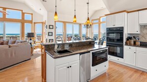 A spacious kitchen with white cabinets, a large island with a sink, and a view of the ocean through large windows. - A Raye Of Sunshine: Ocean Views, Saltwater Pool (Corolla)