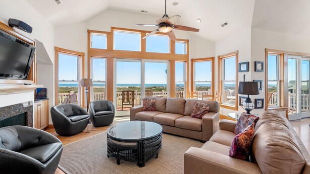 A spacious living room with a large window overlooking the beach, a ceiling fan, and a flat-screen TV mounted on the wall. - A Raye Of Sunshine: Ocean Views, Saltwater Pool (Corolla)