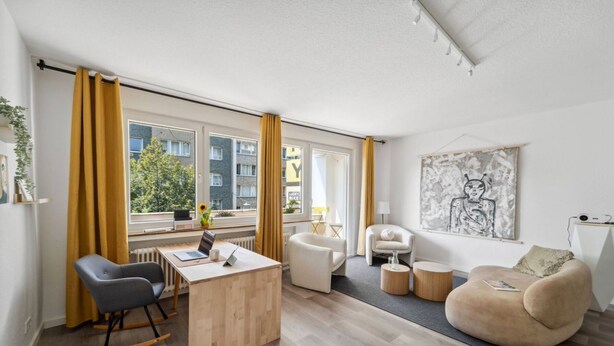 A modern living room with a desk, a sofa, and a chair. There are windows with curtains, a radiator, and a painting on the wall. - Business Flat in Little Tokyo (Düsseldorf)