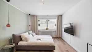 A modern bedroom with a bed, a small side table, a TV mounted on the wall, and a window with curtains. - Business Flat in Little Tokyo (Düsseldorf)