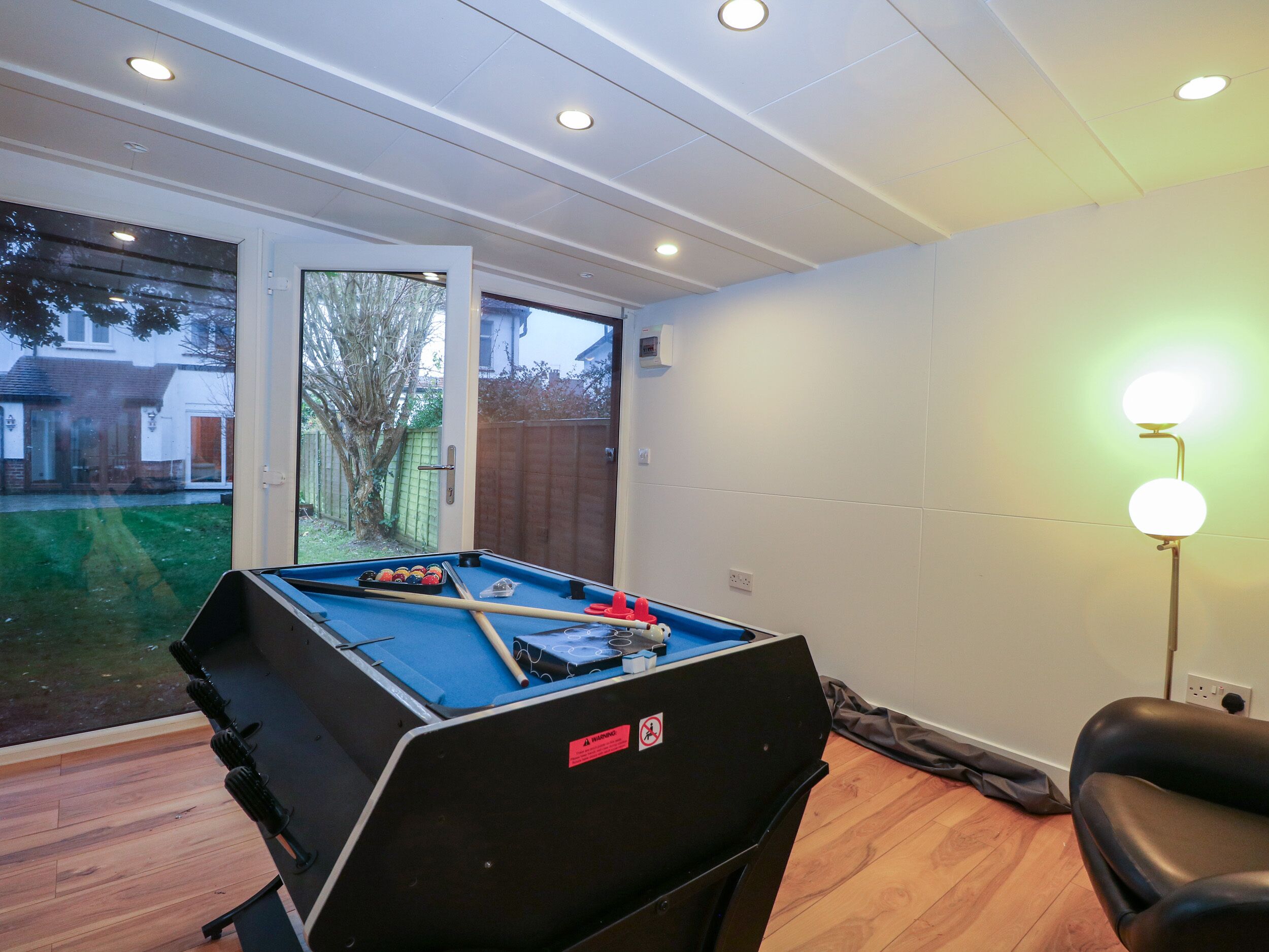 A pool table in a room with a door leading to a backyard, a black chair, and a floor lamp.