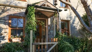 Une porte en bois rustique, intégrée à un portail en bois, le tout entouré d’une végétation luxuriante et d’un mur de pierres.