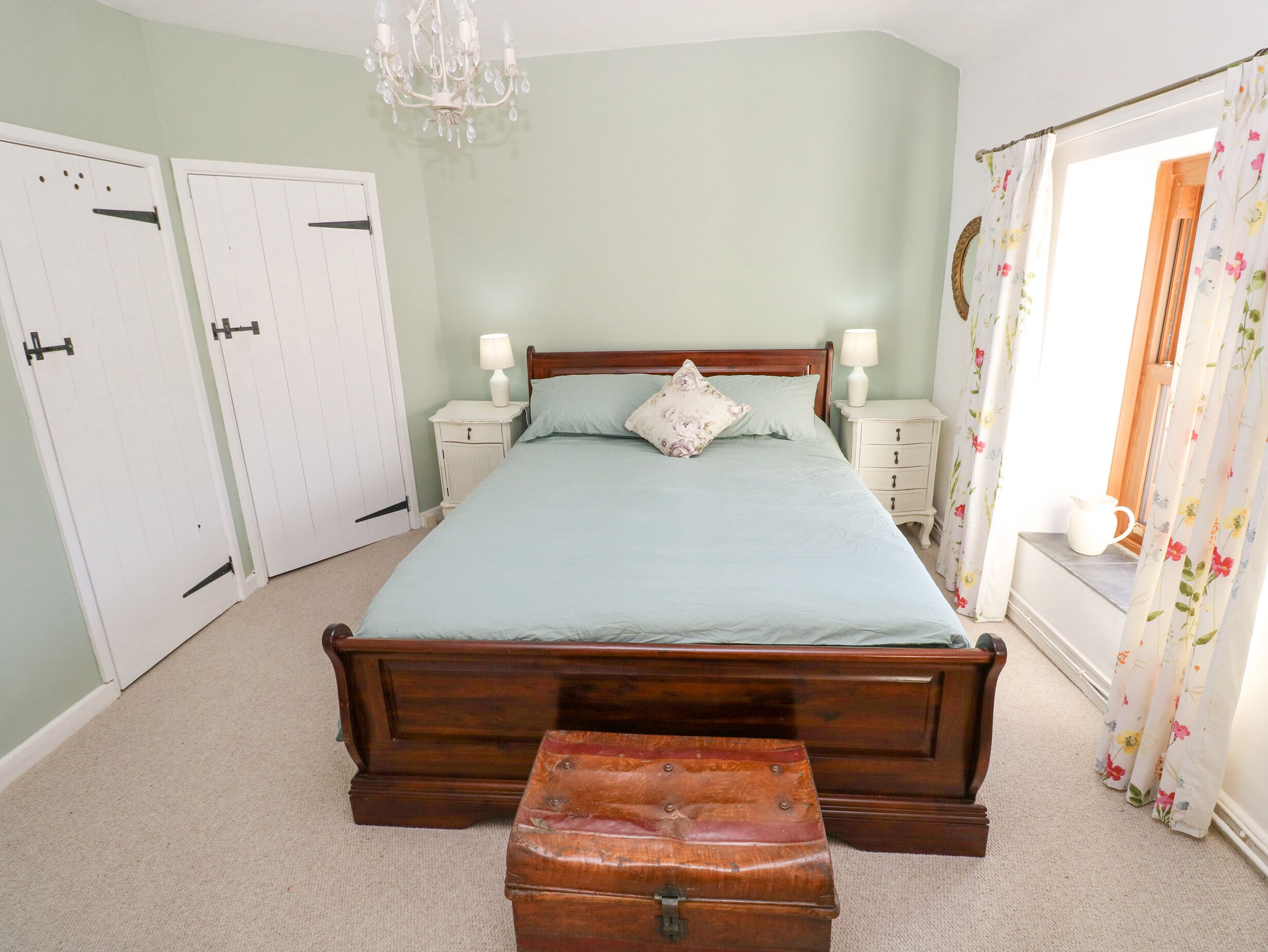 A bedroom with a wooden bed, two bedside tables with lamps, a nightstand, a chest of drawers, a chandelier, and a window with floral curtains.