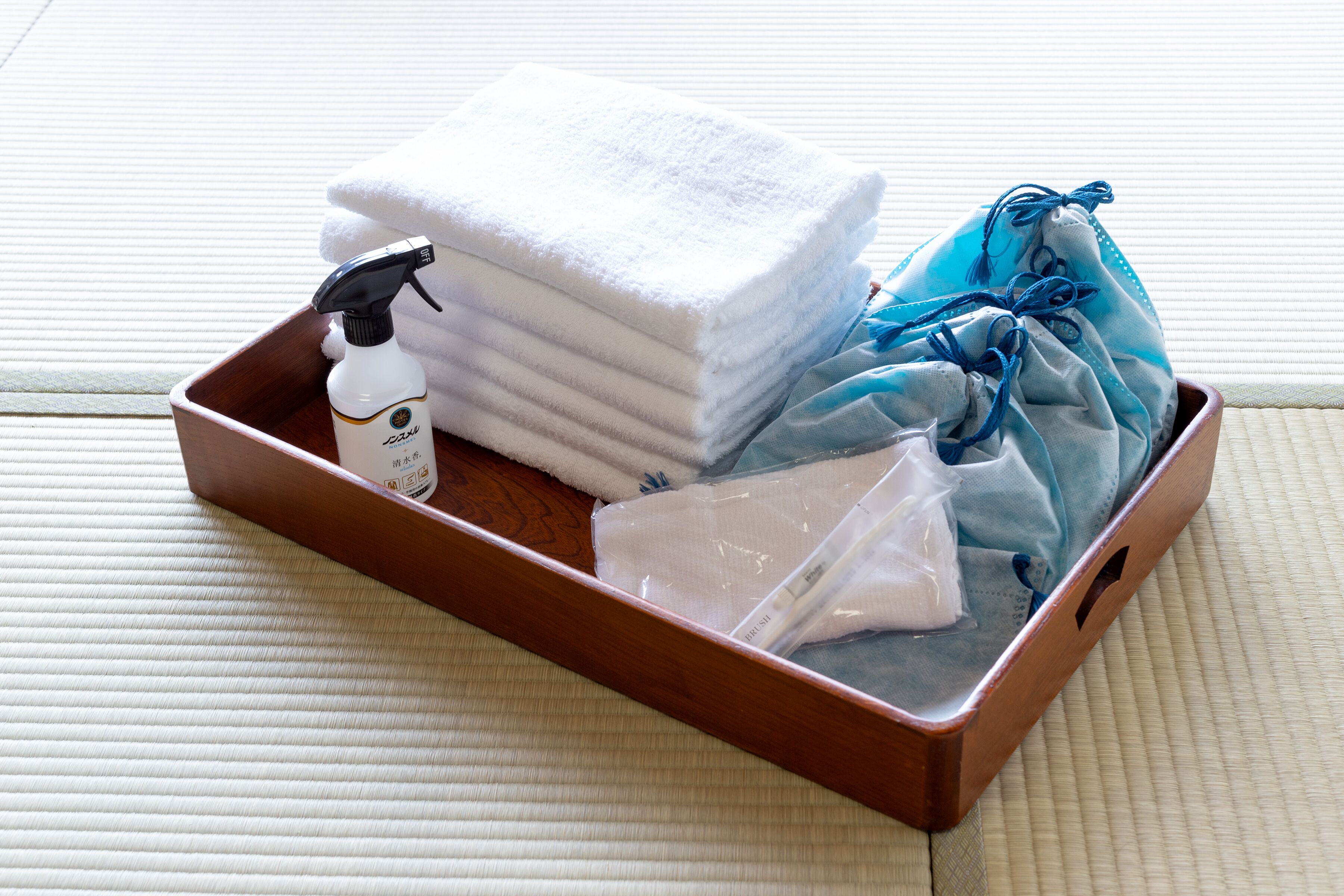 A wooden tray with a bottle of shampoo, folded towels, and a blue cloth on a textured mat.