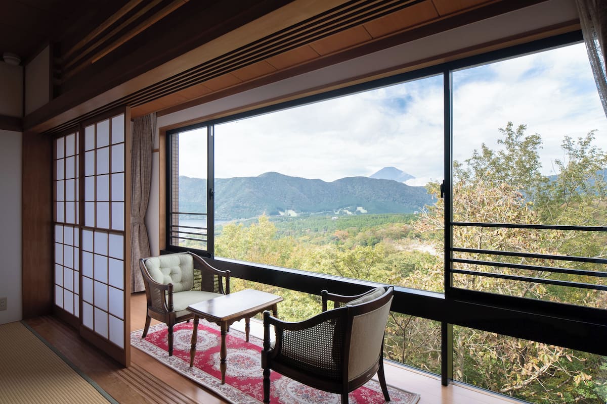 Ein traditioneller japanischer Raum mit Tatami-Boden, einem niedrigen Tisch, zwei Stühlen und Blick auf Berge durch große Fenster.