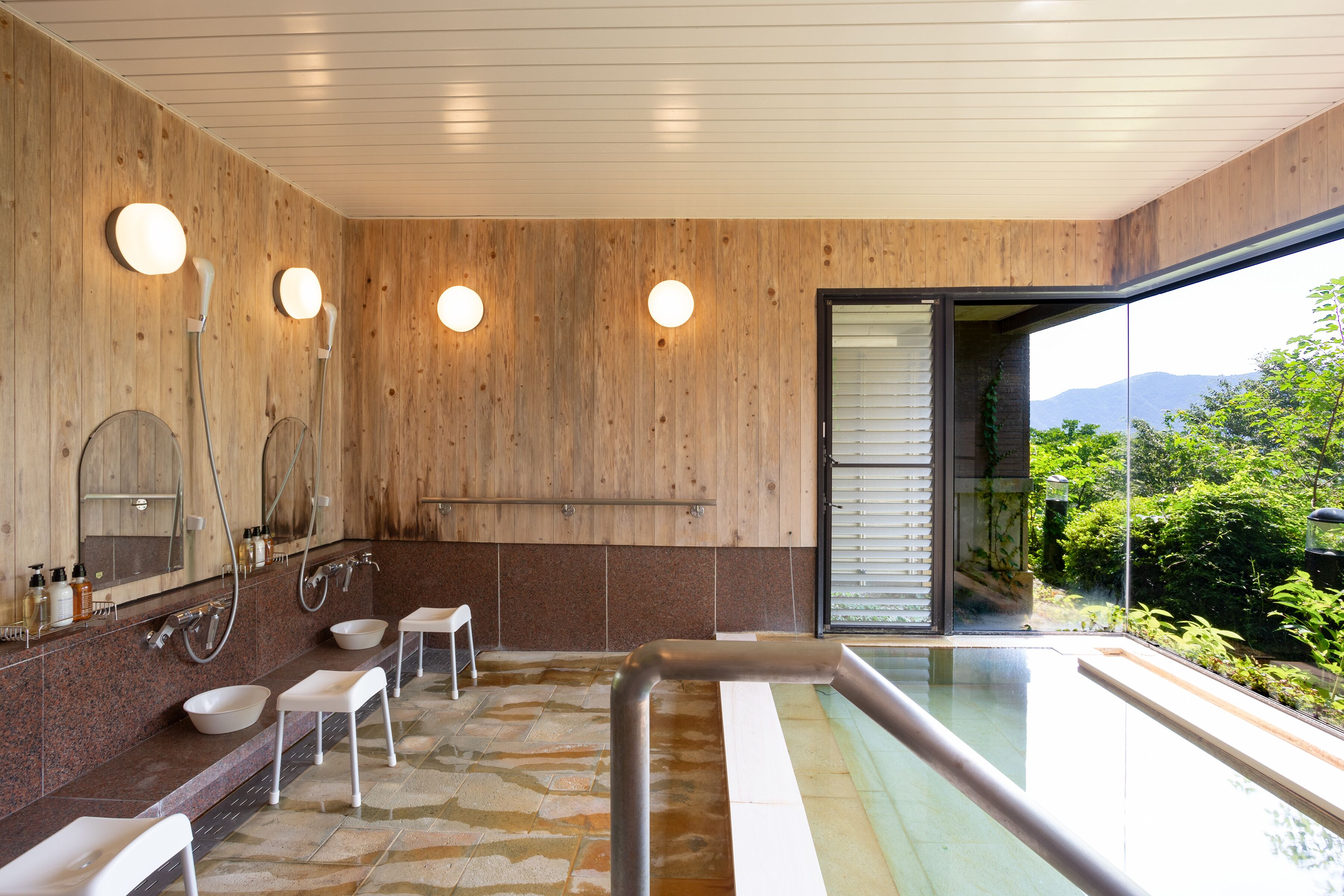 A modern bathroom with wooden walls, a large mirror, a bathtub, and a view of greenery.