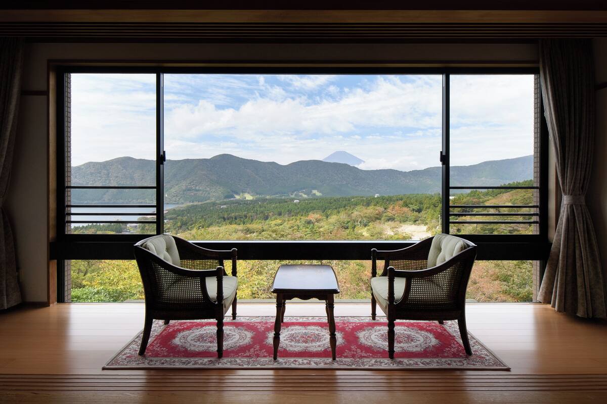 Ein Zimmer mit großem Fenster und Blick auf Berge und Grünflächen. In der Mitte stehen zwei Korbstühle und ein kleiner Tisch.