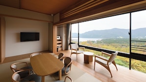 A modern interior with a wooden floor, a dining table, chairs, a TV mounted on the wall, and a large window offering a view of mountains and a lake. - Hakone Ashinoko Bikeikan (Hakone)