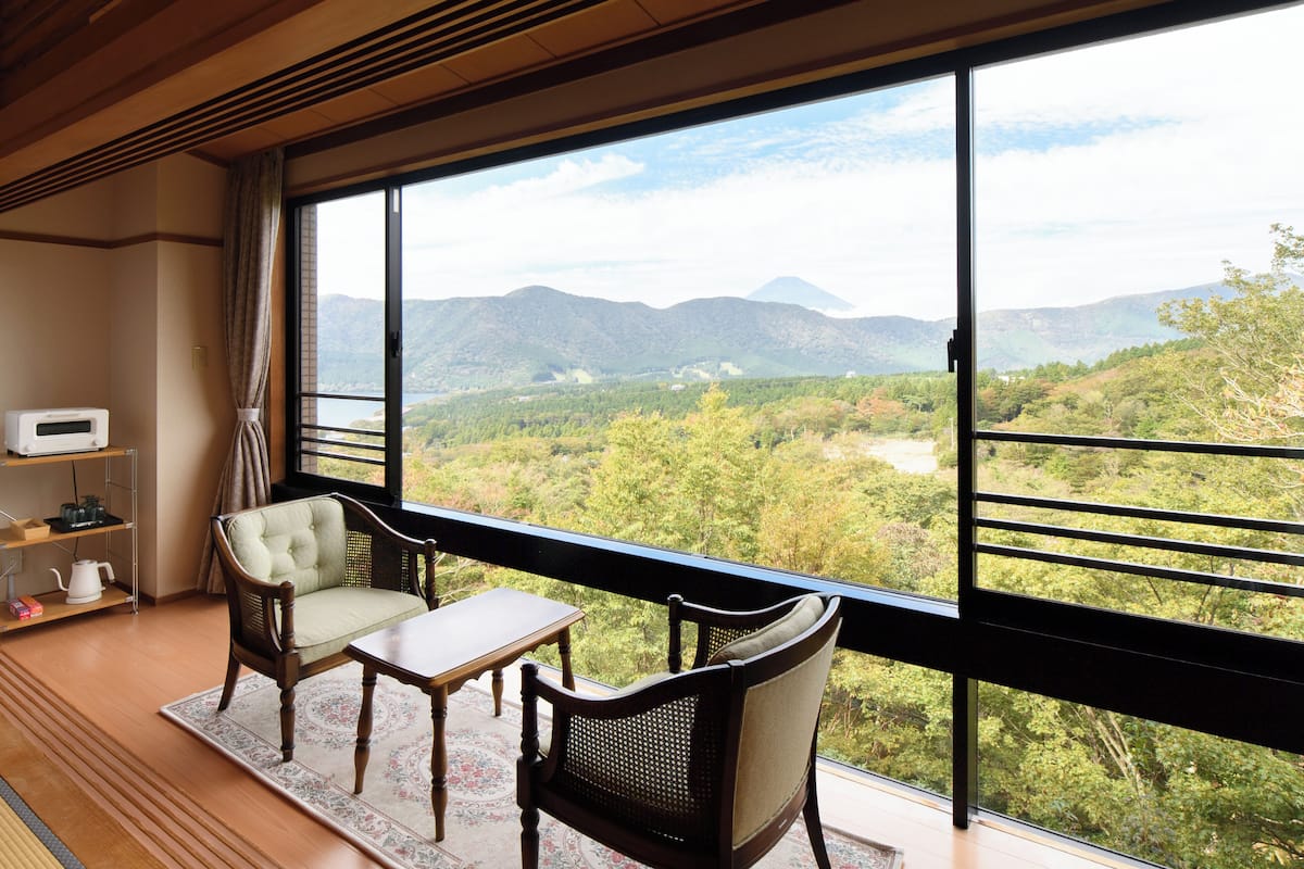 Ein Zimmer mit großem Fenster und Blick auf eine gebirgige Landschaft. Ein Holztisch und zwei Stühle sind vorhanden.