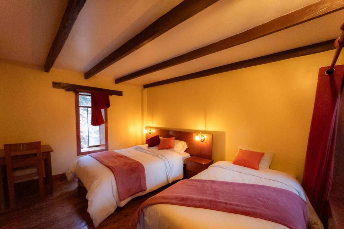 A room with two beds, wooden beams on the ceiling, a window with a view, and a wooden chair.