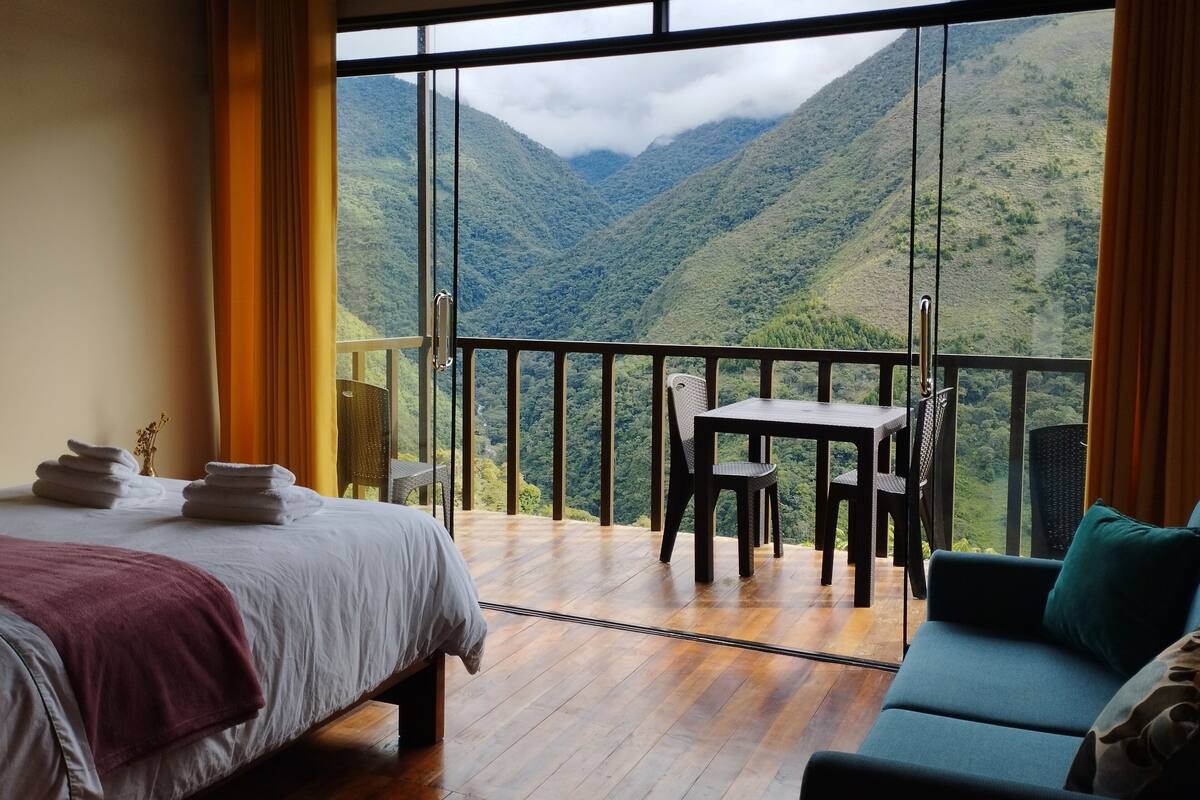 A room with a bed, a table, and a sofa, with a view of a mountainous landscape through large windows.