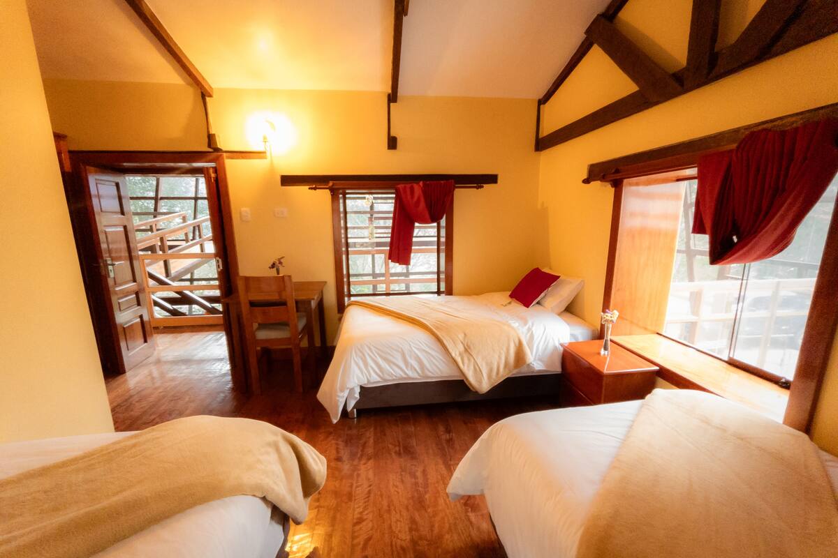 A cozy room with two beds, a desk, and a chair. There is a window with curtains and a lamp on the wall.