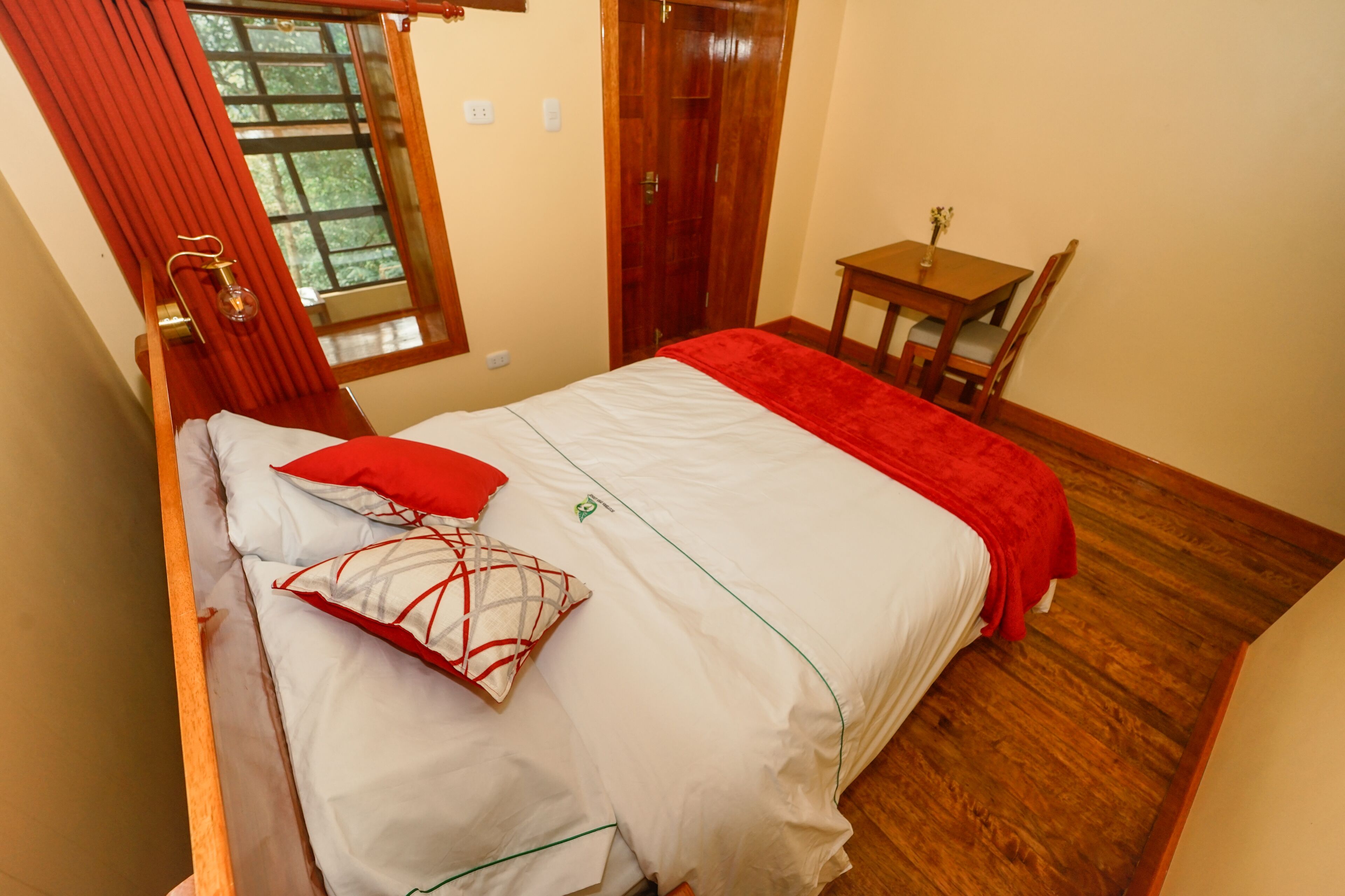 Un lit bien fait, avec une literie blanche et rouge, une chaise en bois, une petite table en bois sur laquelle est posé un vase, et une fenêtre ornée de rideaux rouges.
