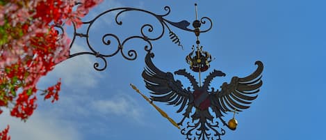 Estrutura metálica decorativa com emblema alado e um sino, ao fundo um céu azul límpido.